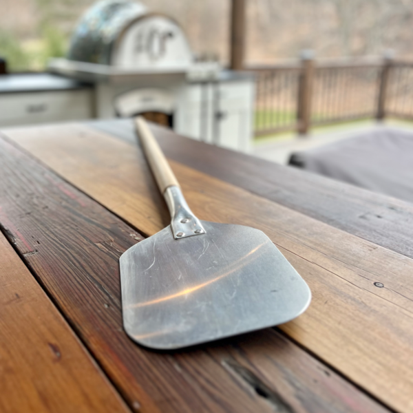 HPC Fire aluminum pizza peel with wood handle displayed on an outdoor kitchen counter beside a wood-fired pizza oven
